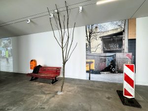 Eine Ausstellungsansicht, die eine Kunstinstallation der Künstlerin Simone Zaugg zeigt. Zu sehen sind eine Bank, ein Ast, ein Bild an der Wand sowie eine Absperrung.