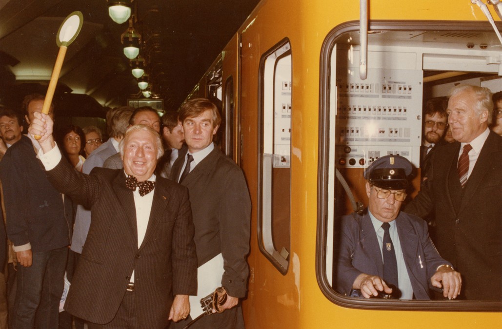 Eine U-Bahn, im Fahrerhäuschen sitzen zwei Personen, links daneben stehen erfreute Personen.